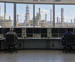 Synthetic editorial illustration. A refinery control room. Two rear-facing operators sit at a curved console of dark display panels and switches, with extensive refinery towers, tanks and pipework visible through broad windows.