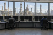 Synthetic editorial illustration. A refinery control room. Two rear-facing operators sit at a curved console of dark display panels and switches, with extensive refinery towers, tanks and pipework visible through broad windows.
