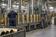 Synthetic editorial illustration. A factory production hall. Empty brass shell casings stand along roller conveyors while two workers in ear protection face away among large industrial machines.
