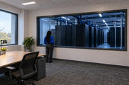 Synthetic editorial illustration. A small rear-facing person with long braids stands beside a broad interior window into a blue-lit server room. A simple desk, chair and plants occupy the quiet adjoining office.