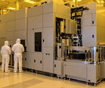 Synthetic editorial illustration. Two small white-suited engineers viewed from behind stand beside a massive plain machine enclosure. Exposed metal mechanisms and a wafer stack sit in a yellow-lit cleanroom, with equipment displays dark.