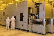 Synthetic editorial illustration. Two small white-suited engineers viewed from behind stand beside a massive plain machine enclosure. Exposed metal mechanisms and a wafer stack sit in a yellow-lit cleanroom, with equipment displays dark.