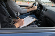 Synthetic editorial illustration. Through an open passenger window, an examiner writes on a populated softly defocused clipboard beside a belted driver holding the steering wheel, with a controlled test area beyond.