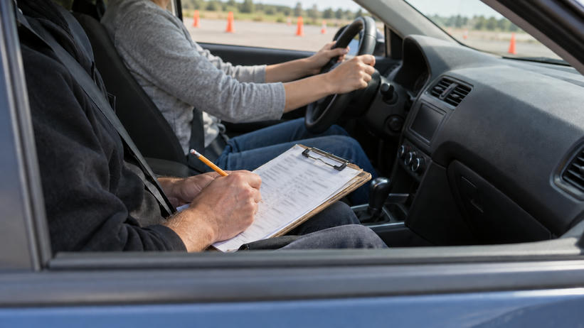 Synthetic editorial illustration. Through an open passenger window, an examiner writes on a populated softly defocused clipboard beside a belted driver holding the steering wheel, with a controlled test area beyond.