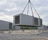 Synthetic editorial illustration. A gravel construction site. A grey modular equipment unit hangs from crane slings above concrete footings, with similar ventilated units already installed nearby.