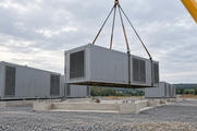 Synthetic editorial illustration. A gravel construction site. A grey modular equipment unit hangs from crane slings above concrete footings, with similar ventilated units already installed nearby.