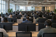 Synthetic editorial illustration. A large open-plan office. Rows of rear-facing workers wearing headsets sit behind plain monitor backs and low desk partitions, with tall windows, plants and exposed ceiling services surrounding the space.