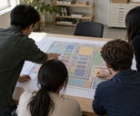 Synthetic editorial illustration. Four people viewed from above and behind gather around a large paper layout of muted coloured blocks and fine geometric connections. Hands point at the sheet on a wooden table in a daylit office.