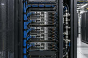 Synthetic editorial illustration. The open mesh door of a cabinet frames installed rack servers, organised network leads and connected power cables. A vertical metal pipe assembly sits beside the rack, with more cabinets receding along the aisle.