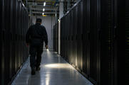 Synthetic editorial illustration. A guard in an unmarked dark uniform walks away along an aisle of black server cabinets. A handheld flashlight casts a small pool of light on the floor beneath sparse ceiling lights and status LEDs.