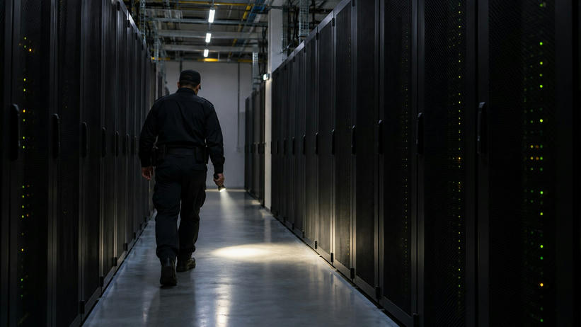 Synthetic editorial illustration. A guard in an unmarked dark uniform walks away along an aisle of black server cabinets. A handheld flashlight casts a small pool of light on the floor beneath sparse ceiling lights and status LEDs.