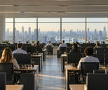 Synthetic editorial illustration. An open-plan office overlooks a broad city skyline through tall windows. Rear-facing analysts sit at rows of desks with dark monitors, plants and chairs arranged along a central aisle.