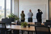 Synthetic editorial illustration. A research office. Three rear-facing colleagues study a whiteboard of empty boxes and arrows, beyond desks with dark monitors, mesh chairs and potted plants in soft daylight.