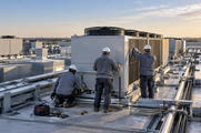 Synthetic editorial illustration. A data-centre rooftop. Three workers in hard hats face a large silver cooling unit, one crouching beside it. Pipes cross the roof and tools lie nearby in warm low sunlight.