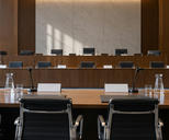 Synthetic editorial illustration. A hearing room contains long wooden tables with microphones, blank placards and water carafes. Most black chairs are empty, with a small rear-facing figure seated at the far left in daylight.