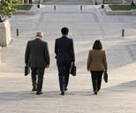 Synthetic editorial illustration. A civic building stairway. Three formally dressed people seen from behind carry document cases down broad stone steps toward a quiet paved plaza.