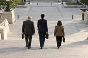 Synthetic editorial illustration. A civic building stairway. Three formally dressed people seen from behind carry document cases down broad stone steps toward a quiet paved plaza.