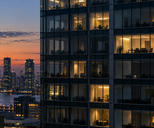 Synthetic editorial illustration. Most windows in a glass office tower are dark, while scattered rooms remain warmly lit. An orange dusk sky and distant city lights fill the background.