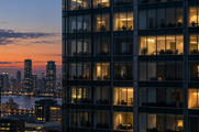 Synthetic editorial illustration. Most windows in a glass office tower are dark, while scattered rooms remain warmly lit. An orange dusk sky and distant city lights fill the background.
