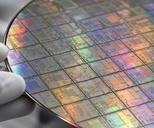 Synthetic editorial illustration. White-gloved fingertips hold the edge of a silicon wafer in extreme close-up. A grid of intricate die patterns reflects muted rainbow colours against a softly blurred laboratory background.