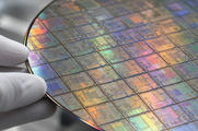 Synthetic editorial illustration. White-gloved fingertips hold the edge of a silicon wafer in extreme close-up. A grid of intricate die patterns reflects muted rainbow colours against a softly blurred laboratory background.