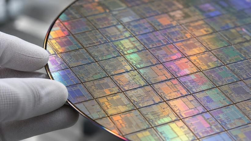 Synthetic editorial illustration. White-gloved fingertips hold the edge of a silicon wafer in extreme close-up. A grid of intricate die patterns reflects muted rainbow colours against a softly blurred laboratory background.