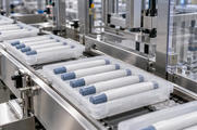 Synthetic editorial illustration. Plain white pen-shaped devices with muted blue-grey caps rest in rows of translucent trays on a stainless conveyor. Metal packaging fixtures and clear protective guards surround the clean production line.