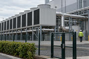 Synthetic editorial illustration. Installed cooling equipment and routed pipework sit beside a secured data-facility perimeter with a small service worker.