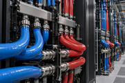 Synthetic editorial illustration. A data-centre hall contains red and blue liquid-cooling hoses connected to metal rack manifolds. Black server cabinets recede along a brightly lit aisle.