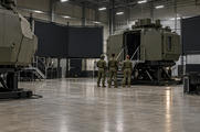 Synthetic editorial illustration. A military simulator hall. Three soldiers seen from behind stand beside elevated armoured cabin mock-ups, with large dark projection screens and an open concrete floor.