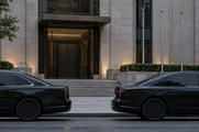 Synthetic editorial illustration. An institutional building entrance. Two dark sedans with opaque windows frame broad steps leading to a plain stone doorway, beside a row of empty flagpoles and clipped hedges.