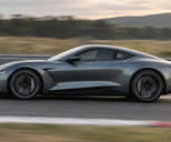 Synthetic editorial illustration. A vehicle test track. A low grey coupe travels past the camera with blurred wheels, striped curbing and a streaked guardrail, beneath a softly lit sky and distant hills.