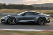 Synthetic editorial illustration. A vehicle test track. A low grey coupe travels past the camera with blurred wheels, striped curbing and a streaked guardrail, beneath a softly lit sky and distant hills.