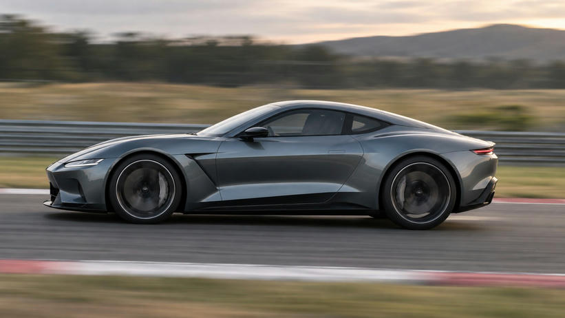 Synthetic editorial illustration. A vehicle test track. A low grey coupe travels past the camera with blurred wheels, striped curbing and a streaked guardrail, beneath a softly lit sky and distant hills.
