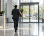 Synthetic editorial illustration. A suited person seen from behind carries a plain cardboard box containing a potted plant through a bright glass office lobby. The polished floor reflects daylight beside empty chairs and a reception counter.