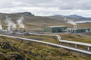 Synthetic editorial illustration. Generic geothermal generating buildings, insulated steam pipes and modest ordinary water vapour in an anonymous upland landscape.