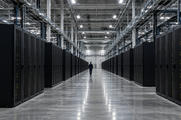 Synthetic editorial illustration. A data-centre hall contains long rows of black equipment racks. Tiny green and amber lights flank a broad polished aisle where a small rear-facing worker walks beneath cable trays.
