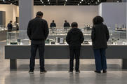Synthetic editorial illustration. A technology exhibition hall. Visitors seen from behind examine circuit boards and metal heat sinks under glass on a demonstration table, with more display cases across the room.