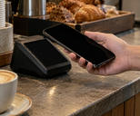 Synthetic editorial illustration. A cafe counter. A cropped hand holds a dark phone beside a plain payment terminal with a black screen, surrounded by a coffee cup, stacked saucers and pastries.