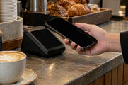 Synthetic editorial illustration. A cafe counter. A cropped hand holds a dark phone beside a plain payment terminal with a black screen, surrounded by a coffee cup, stacked saucers and pastries.