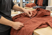 Synthetic editorial illustration. Hands check the zipper of one unbranded technical jacket at a sportswear retail counter.