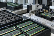 Synthetic editorial illustration. Generic packaged integrated circuits and memory boards in anti-static trays beside an electrical test fixture. Chip assembly/packaging and testing, not wafer fabrication or laptop assembly. No actual named company, product, site, person or event is depicted.