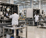 Synthetic editorial illustration. Three technicians viewed from behind work on large open precision-equipment modules with mounted mirrors. Metal stands, component trolleys and plain blue storage bins fill a brightly lit assembly hall.