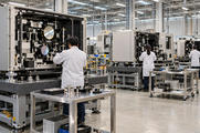 Synthetic editorial illustration. Three technicians viewed from behind work on large open precision-equipment modules with mounted mirrors. Metal stands, component trolleys and plain blue storage bins fill a brightly lit assembly hall.