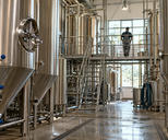 Synthetic editorial illustration. Stainless fermentation tanks and hygienic pipework in an anonymous brewery, with a distant worker seen from behind.
