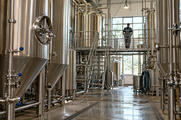 Synthetic editorial illustration. Stainless fermentation tanks and hygienic pipework in an anonymous brewery, with a distant worker seen from behind.