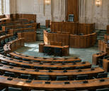 Synthetic editorial illustration. A parliamentary chamber contains curved wooden benches and green upholstered chairs. Desk microphones face a raised speaker's chair, with soft daylight falling across the empty seating.