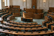 Synthetic editorial illustration. A parliamentary chamber contains curved wooden benches and green upholstered chairs. Desk microphones face a raised speaker's chair, with soft daylight falling across the empty seating.
