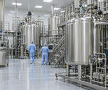 Synthetic editorial illustration. A biologics production hall. Two operators in pale-blue gowns seen from behind stand among tall stainless vessels, dense pipework, processing skids and dark control screens.
