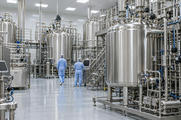 Synthetic editorial illustration. A biologics production hall. Two operators in pale-blue gowns seen from behind stand among tall stainless vessels, dense pipework, processing skids and dark control screens.